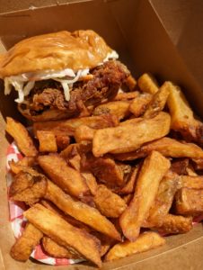 Fried chicken sandwich and thick-cut French fries in a cardboard takeout container - All Out Burger