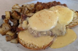 The sausage eggs Benedict - a toasted English muffin topped with a patty of the house-made sausage and two poached eggs, all slathered in a muted-yellow hollandaise sauce and a sprinkling of red paprika, with sides of grilled home fries and baked beans in the background. 