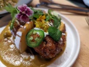 A close up shot of the pork belly pogo - the pogo is colourfully topped with cilantro, fresh jalapeno and peach, and cute little edible flowers, while sitting in a green tomato salsa - Fraser Restaurant