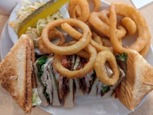 Classic club sandwich, cut into four triangle-shaped quarters, with onion rings spilling over onto the sandwich from their side of the plate, with a side dish of coleslaw and a pickle spear.