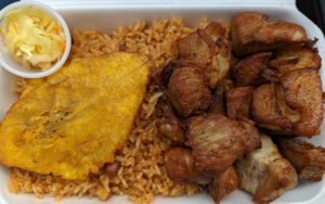 Griot plate - chunks of seasoned and fried pork, on a bed of rice and beans, with a large, flattened fried plantain, with a little cup of pikliz slaw in the corner. Margo Xpress