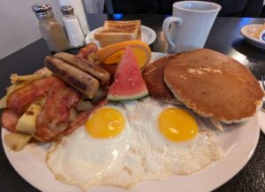 Fontenelle's Ultimate Breakfast Platter - a large, white, oval plate is piled with food - two sunny-side-up eggs in the middle are flanked by two pieces each of bacon and breakfast sausage which are on top of griddled, house-made homefries. Two pancakes are on the right side. A small wedge of watermelon and a slice of orange are the lone items of colour and nutrition. A plate with two pieces of toast, which presumably couldn't fit on the main plate, is in the background, with a plain white mug of coffee beside it.