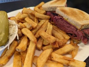 Fontenelle - smoked meat sandwich - a large white plate has a pile of fries in the middle, with thick stack of slices of Montreal smoked meat in two halves of a sandwich, between two toasted slices of white bread. A small bowl of coleslaw, with a pickle on top, sits on the left edge of the plate.